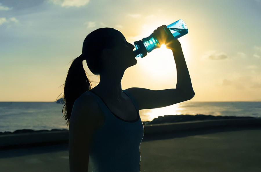 A woman in exercise clothes drinking from a water bottle on the beach, with the water and sunset in the background. Get EDATATE detoxification treatment from Balanced Medical Solutions in San Francisco Bay Area.