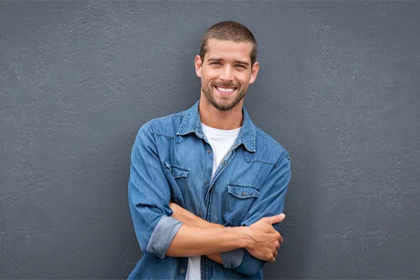 A man in a denim shirt stands smiling against a gray-blue wall, pleased with his testosterone hormone therapy from Balanced Medical Solutions in San Francisco Bay Area.