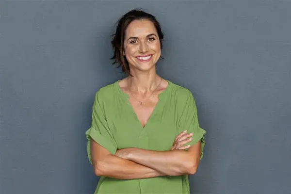A woman in a green shirt stands smiling against a gray-blue wall, pleased with her hormone therapy from Balanced Medical Solutions in San Francisco Bay Area.