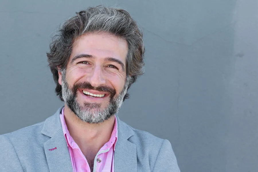 A smiling man with salt and pepper hair and beard wearing a pink shirt and gray blazer stands agains a gray wall. Get hypogonadism treatment from Balanced Medical Solutions in San Francisco Bay Area.