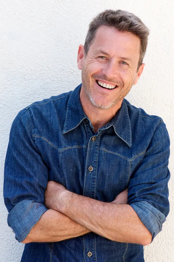 A healthy middle-aged man in a denim shirt leans against a white wall smiling. Get Ipamorelin peptide treatment from Balanced Medical Solutions in San Francisco Bay Area.