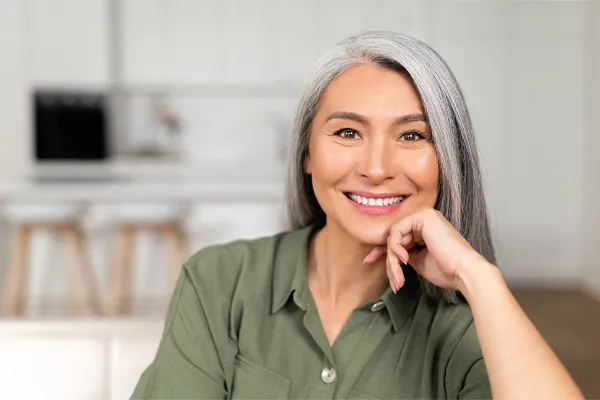 Menopause-Clinic An asian woman with long gray hair and a green shirt sitting and smiling on her couch, happy with her menopause care from [NAME} in San Francisco Bay Area.