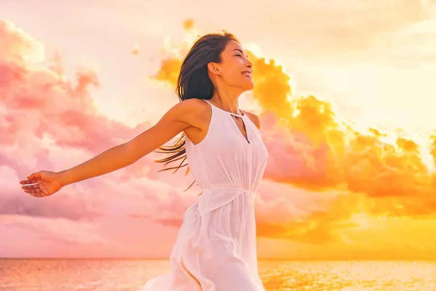 A woman in a thin white dress stretches out her arms joyously on the beach at sunrise. Get the Myers Cocktail IV treatment from Balanced Medical Solutions in San Francisco Bay Area.