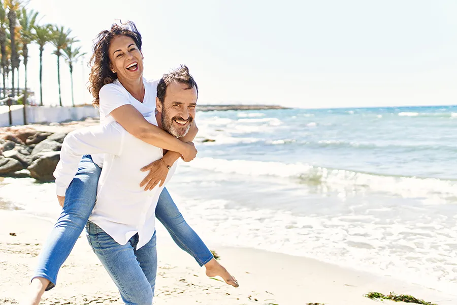 NAD-Therapy-Clinic A woman piggybacks on her husband as the middle-aged couple have fun at the beach. Get NAD Therapy from Balanced Medical Solutions in San Francisco Bay Area.