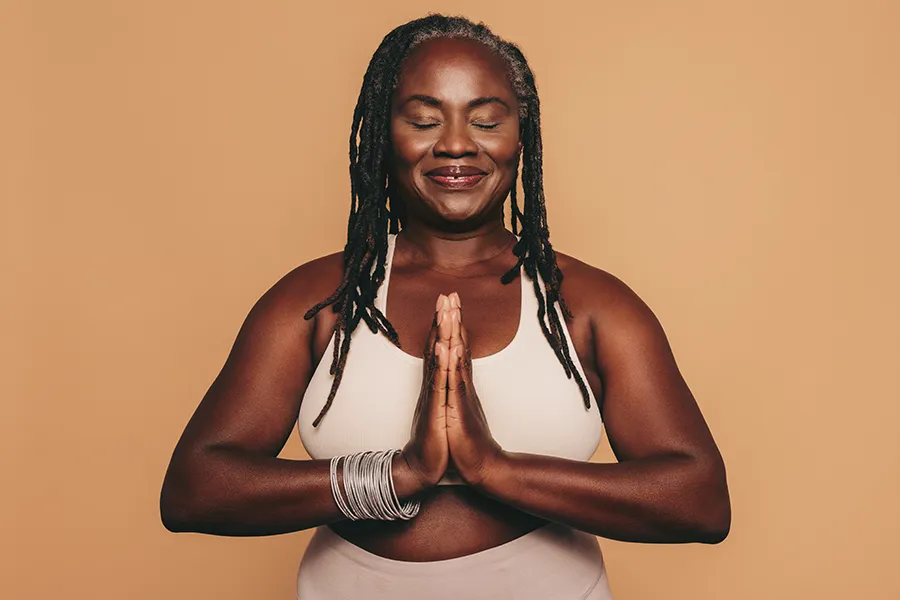 Obesity-Clinic An older African American woman confidently posing against a warm background smiling with hands in a prayer pose. Balanced Medical Solutions treats obesity using functional medicine in San Francisco Bay Area.