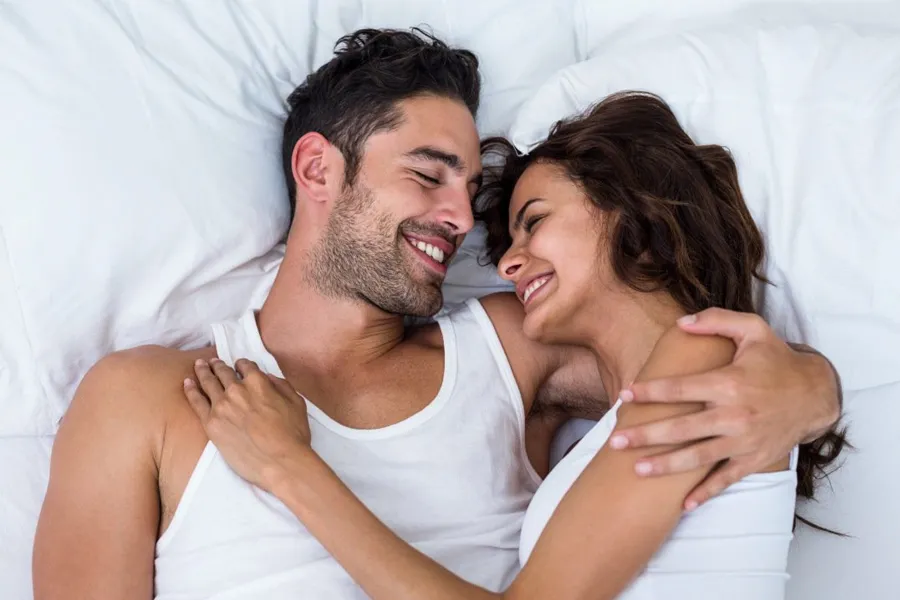 A laughing man and woman in undershirts lie in each other's arms on a white bed, representing successful PT141 sexual wellness therapy for men from Balanced Medical Solutions in San Francisco Bay Area.