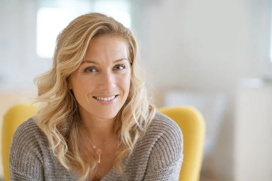 A blonde woman wearing a a gray sweater sits inside in a yellow chair, smiling and happy with her perimenopause treatment from Balanced Medical Solutions in San Francisco Bay Area.