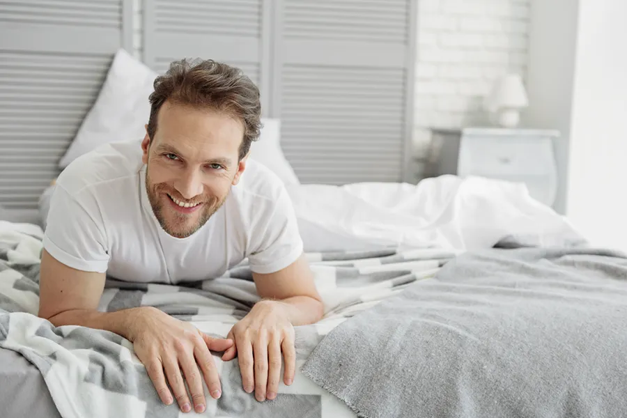 A man in a t-shirt lies on his stomach in bed smiling, happy with his sexual wellness treatments from Balanced Medical Solutions in San Francisco Bay Area.