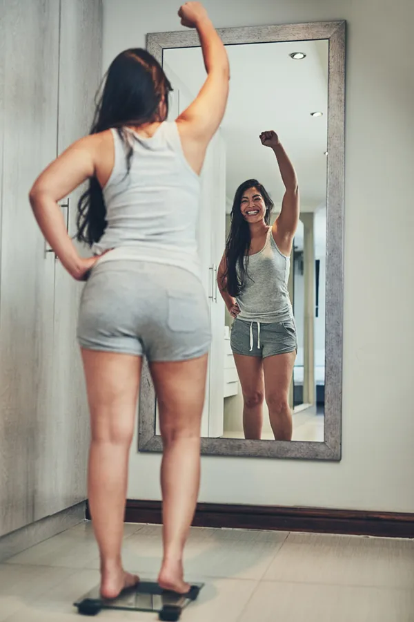 A woman in workout clothes stands on a scale looking in the mirror and pumping her hands in celebration of her successful Tirzepatide treatment from Balanced Medical Solutions in San Francisco Bay Area.